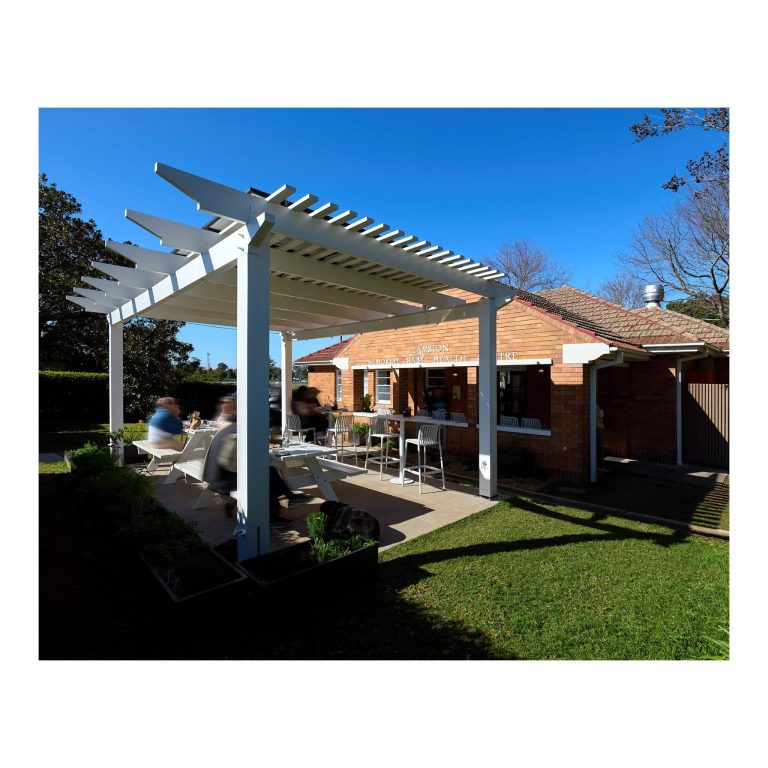 Angle view of a white timber outdoor dining pergola with patrons seated at picnic tables beside the historic red-brick Lambton Baby Health Centre building after heritage building certification.