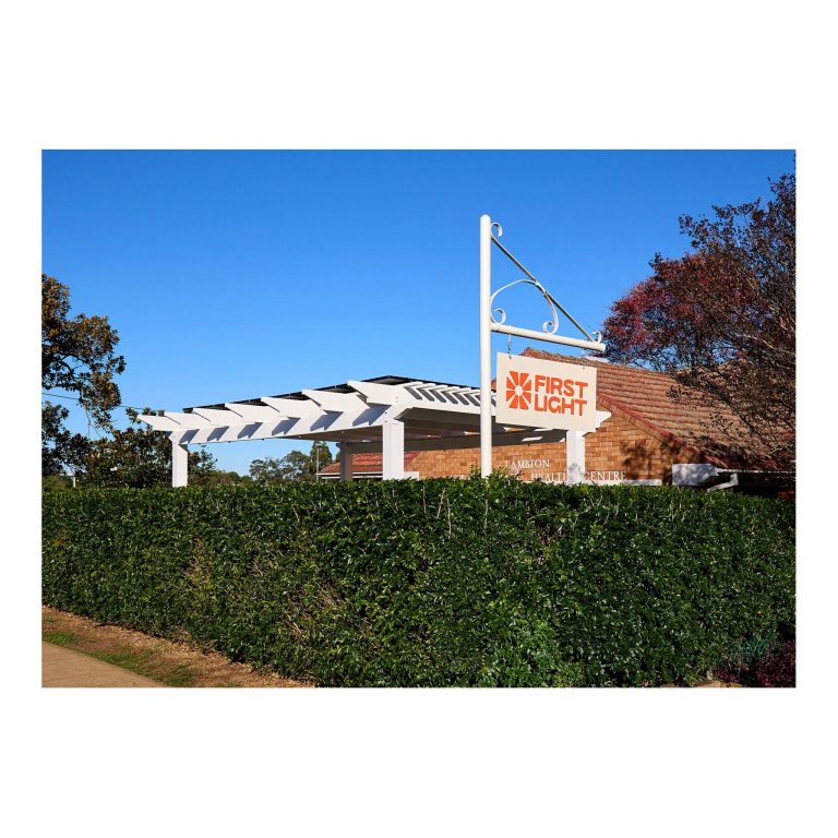 White modern pergola frame structure above a lush green hedge with a white and orange sign reading First Light mounted above a brick building.
