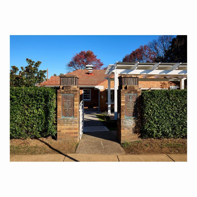 Heritage brick entry pillars with WWII memorial bronze plaques framing a white gate and paved pathway leading to a brick building under a white pergola.