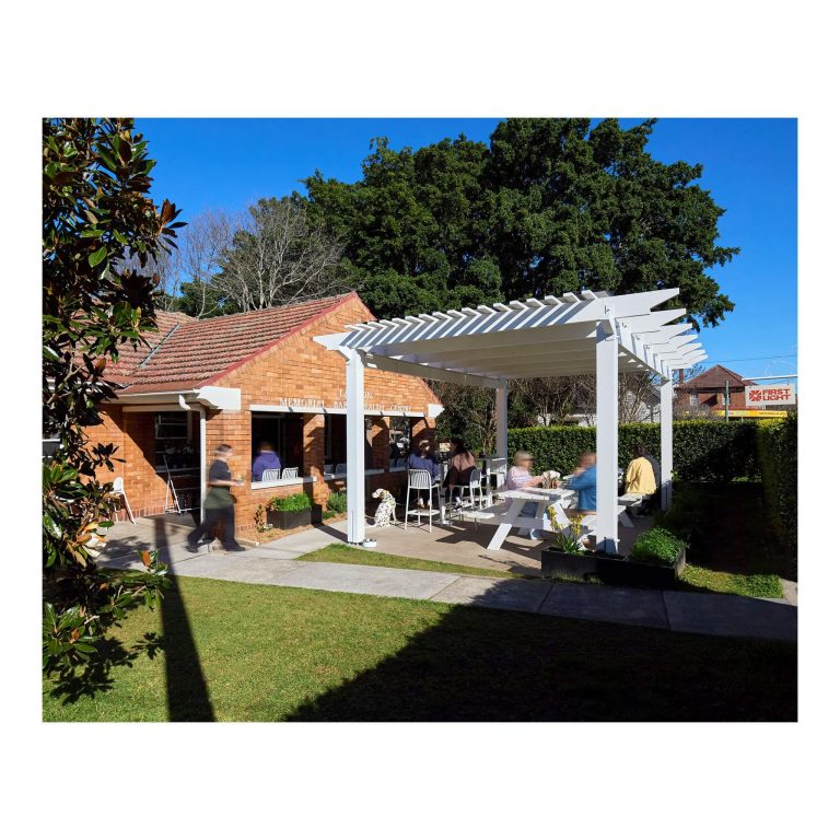 Sunlit outdoor cafe patio with a white wooden pergola structure over picnic tables where patrons sit near a heritage red brick building all inside the hedge.