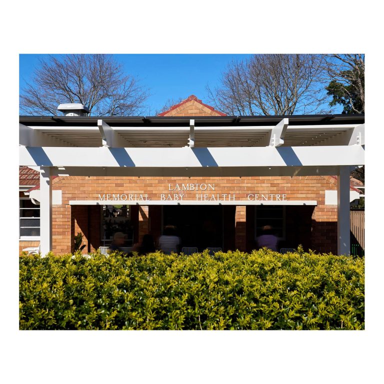 Straight-on front view of the heritage red brick Lambton Memorial Baby Health Centre signage on the building in Lambton NSW, featuring white modern pergola beams above a hedge, with the original heritage building name clearly visible.