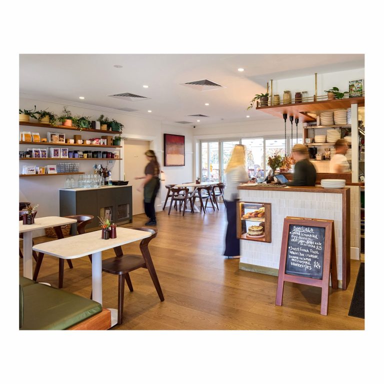 Interior view of First Light cafe showing wooden floors, dining tables, a serving counter with a chalkboard menu, and staff working in the background kitchen area.