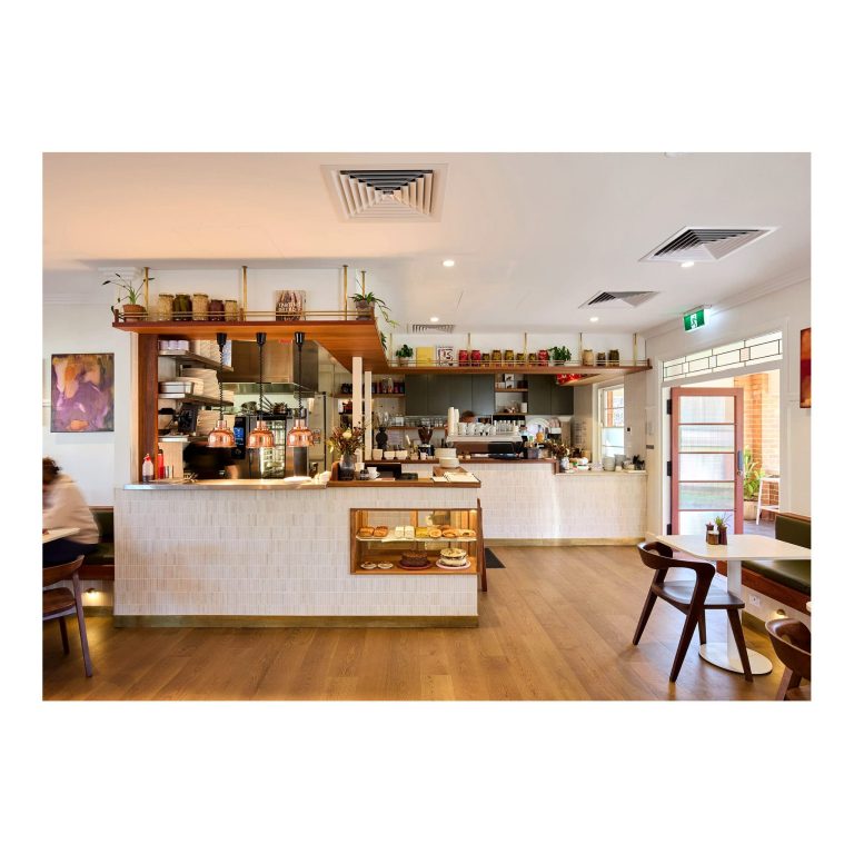 Bright indoor service counter in a modern cafe featuring warm pendant lights, glass display case with baked goods, tiled counters, and timber flooring.
