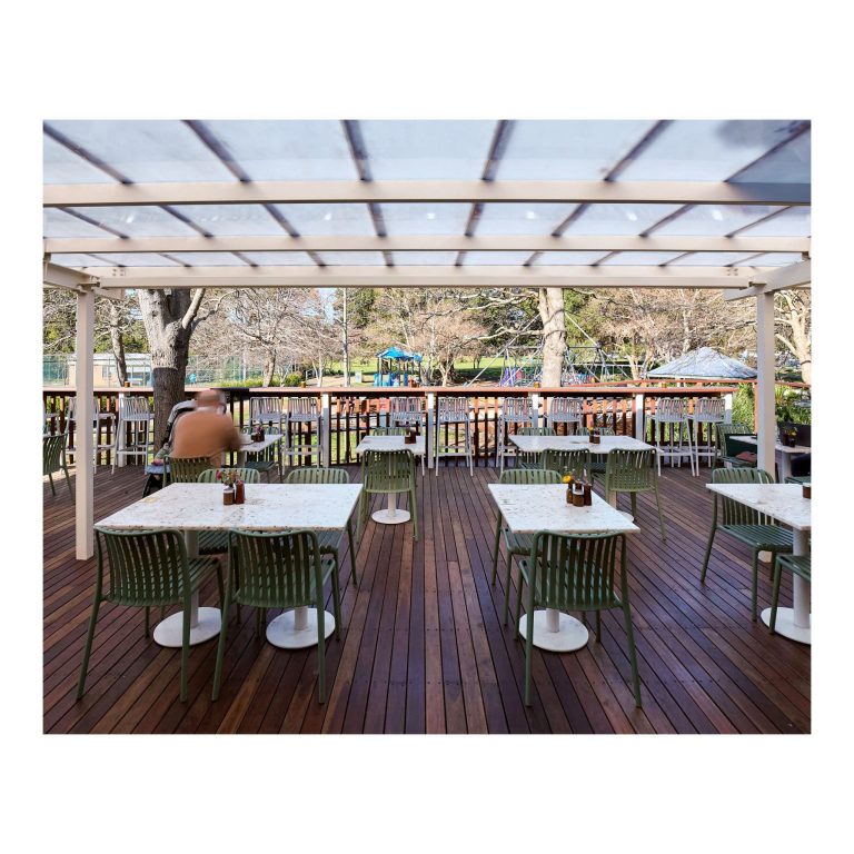 View looking across the outdoor wooden deck at Lambton Park, showing green metal chairs and white speckled dining tables beneath a clear-roofed white pergola, with the park playground in the background.