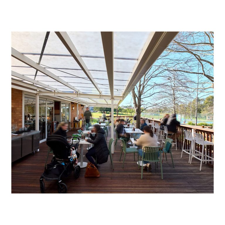 Patrons seated at outdoor tables on the timber deck of First Light Lambton, featuring bi-fold glass doors, a covered pergola, and a view of the surrounding park grounds.