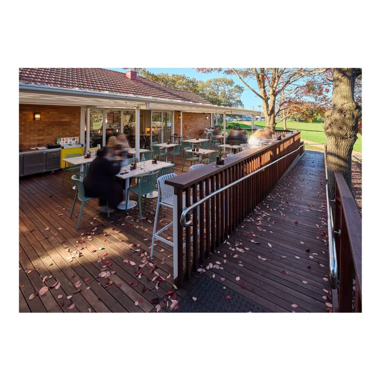 Wide timber outdoor deck with built-in accessibility ramp and handrails, featuring patrons seated at tables alongside a red-brick building with bi-fold doors.