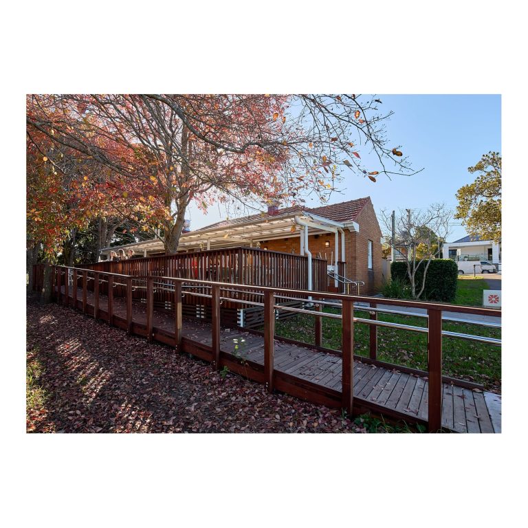 Long timber wheelchair accessibility ramp with stainless steel handrails leading alongside a certified red brick heritage building under autumn trees.