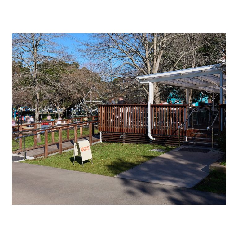 White A-frame sign with orange First Light logo standing in front of timber outdoor deck steps with stainless steel handrails and covered pergola.