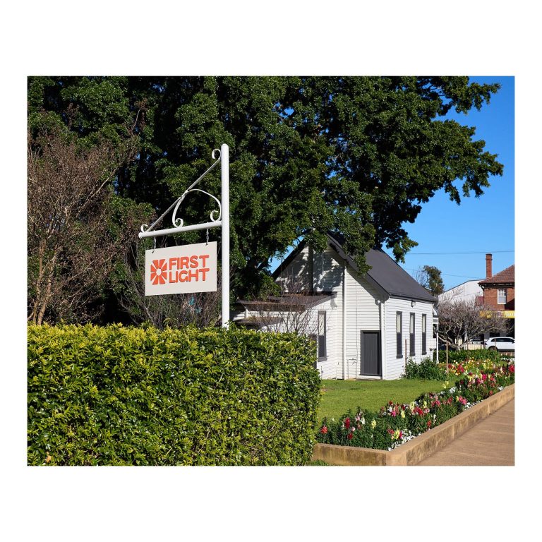 White First Light sign with an orange logo mounted on a post above a green hedge, with the historic weatherboard Lambton Library building and flower beds in the background.
