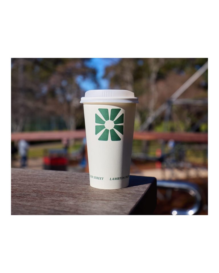 Takeaway coffee cup with a green starburst emblem sitting on a wooden bench, with the blurred playground equipment of Lambton Park visible in the background.