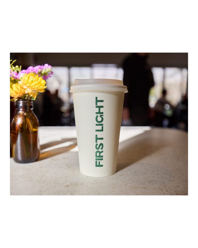 Takeaway coffee cup with vertical green text reading "FIRST LIGHT" standing on a light-colored indoor table next to a small amber glass bottle with yellow and purple flowers.