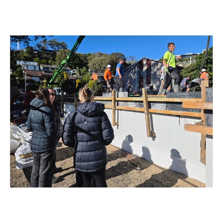 Dominique, Holly, and Tahlia from Buildingwise standing with backs to the camera, on-site team training, observing an active concrete pour and timber formwork on a residential site in Merewether NSW.