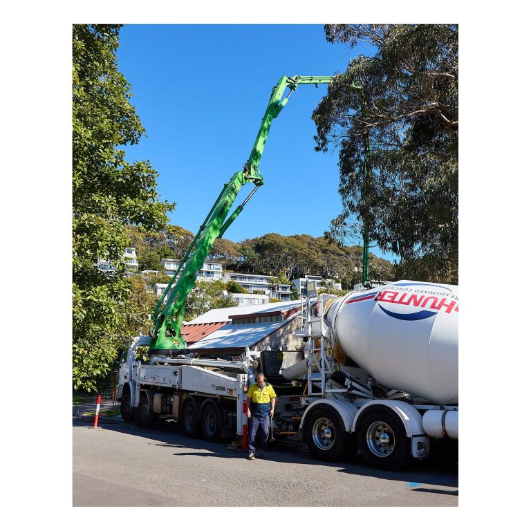 Large white Hunter Readymixed Concrete truck feeding liquid concrete into a tall green Schwing concrete boom pump parked along a residential street in Merewether NSW.