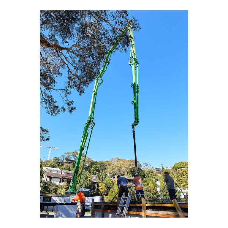High-angle shot of a bright green concrete boom pump extended high above the tree canopy over a residential building site in Merewether NSW.