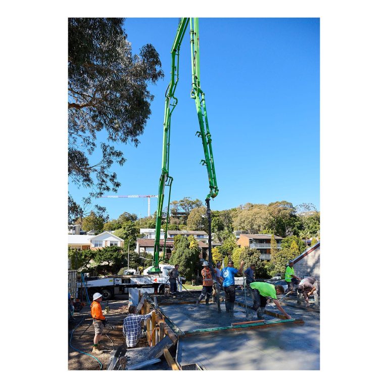 Wide view of multiple concreters working on a large concrete slab pour with a green boom pump elevated against a blue sky in Merewether.