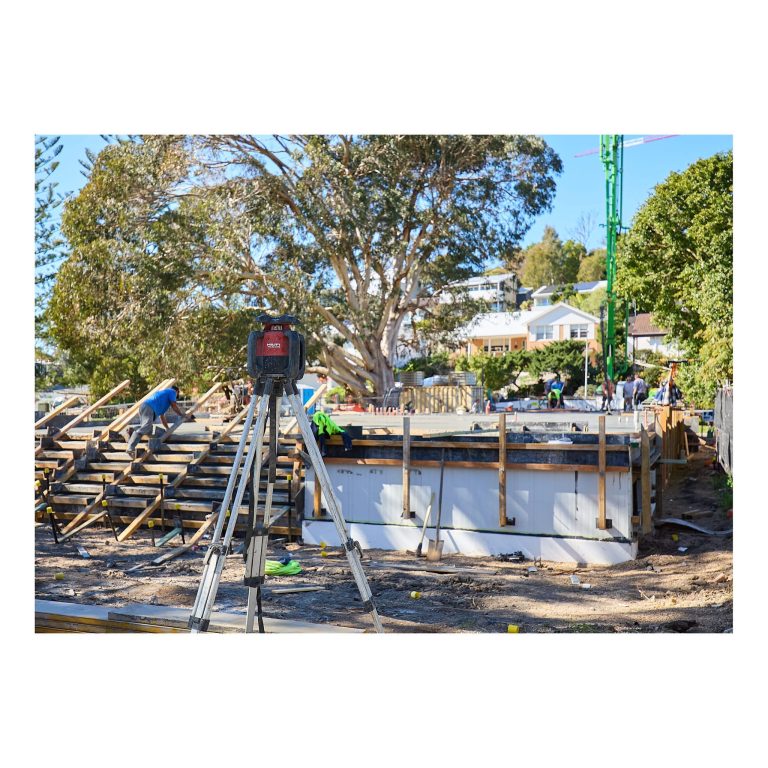 A red Hilti laser level on a tripod set up in the foreground, with workers levelling a concrete slab in the background on a Merewether building site.