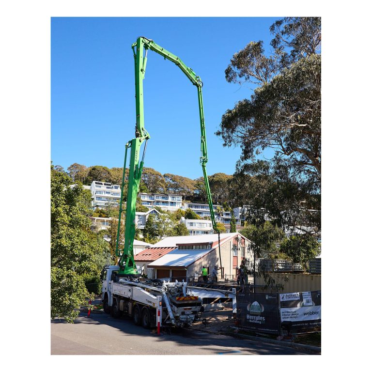 Large white concrete pump truck with a fully extended green boom arm operating on a street in Merewether NSW, with Hercules Building and Buildingwise Certifiers signage on the site fence.