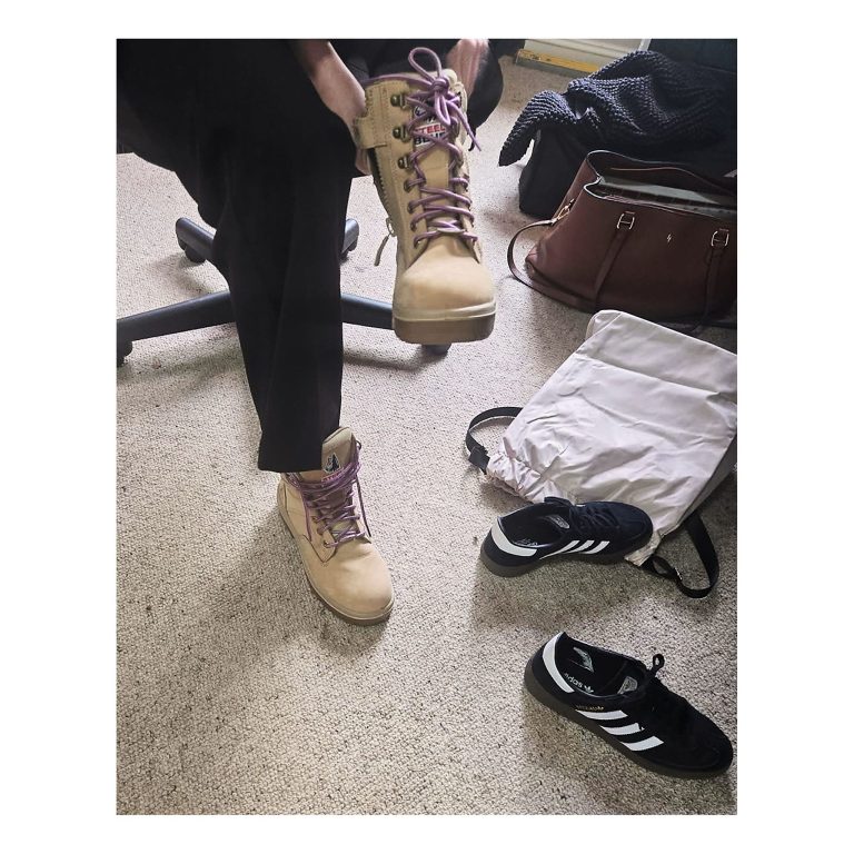 High-angle view of a team member lacing up beige Steel Blue safety work boots with purple laces, preparing for a site inspection, with Adidas sneakers nearby.