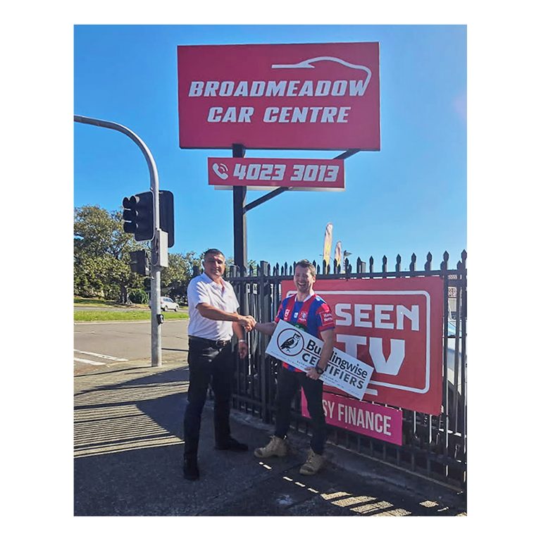 Roby from Broadmeadow Car Centre shaking hands with Kurt from Buildingwise Certifiers, who is wearing a red and blue Newcastle Knights NRL jersey and holding a company sign in front of a red dealership sign and fence banner in Broadmeadow, NSW.